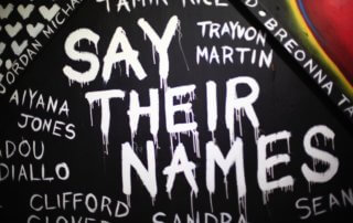 Say Their Names spray painted in white on a dark wall also with names of black lives that have been murdered by police
